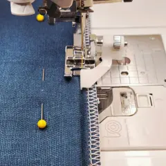 A sewing machine with a blind stitch foot is about to hem dark blue fabric. Pins hold the fabric in place.