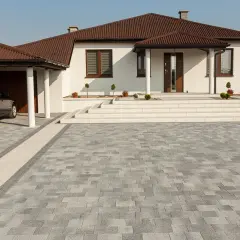 Nowy podjazd z kostki brukowej, z uwzględnieniem jej wymiarów, prezentuje się elegancko obok białego domu z brązowym dachem.