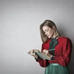Młoda kobieta w okularach i czerwonej bluzce z zieloną kokardą pochyla się nad książką, notując coś ołówkiem. To inspiracja, jak napisać esej historyczny.