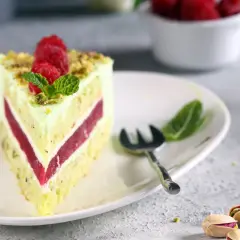 Pyszny tort pistacjowo malinowy z malinami i miętą na talerzu. Obok widoczne są pistacje.