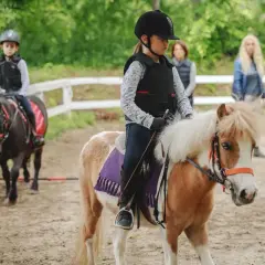 Dzieci w kaskach i kamizelkach ochronnych, w pełnym stroju do jazdy konnej, dosiadają kucyków na piaskowej ujeżdżalni.