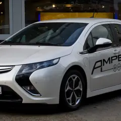 Biała Opel Ampera z czarnym napisem "AMPERA" na drzwiach, zaparkowana przed salonem samochodowym.