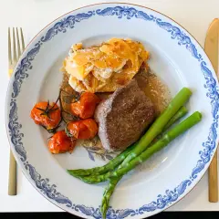 En tallrik med oxfilé sous vide, gratinerad potatis, körsbärstomater och sparris. Guldfärgad kniv och gaffel.