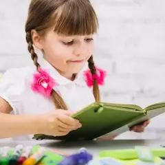 Dziewczynka z warkoczami i różowymi kokardkami pochłonięta lekturą. Jej przygotowanie pedagogiczne widać w skupieniu na nauce.