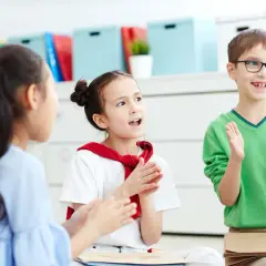 Dzieci na zajęciach wokalnych śpiewają i klaszczą. Chłopiec w okularach trzyma kartkę z literą "LI".