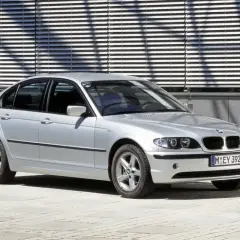 Srebrne BMW E46 sedan z charakterystycznymi podwójnymi reflektorami i grillem, zaparkowane przed nowoczesnym budynkiem z żaluzjami.