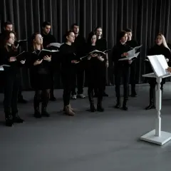 Chór śpiewa, a dyrygentka stoi przy pulpicie. Wszyscy ubrani na czarno, tworząc harmonijną całość, wyższy od basu.