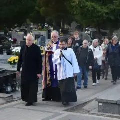 Ksiądz niesie urnę na pogrzebie, a za nim idzie grupa żałobników przez cmentarz.