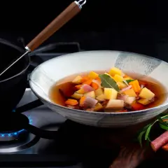 En skål med klar grönsakssoppa med potatis, morötter och lök. En kastrull med soppa värms på spisen.