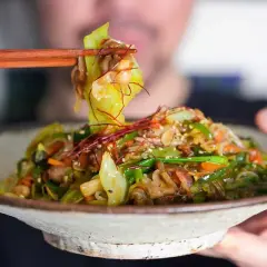 A person uses chopsticks to lift a bite of a colorful Japanese stir fry, featuring cabbage, carrots, and peppers, from a rustic bowl.