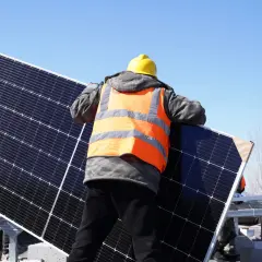 Pracownik w kamizelce odblaskowej i kasku montuje panele fotowoltaiczne. Trwa projekt instalacji fotowoltaicznej.