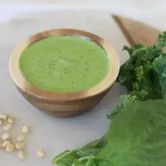 Un cuenco de madera contiene un vibrante pesto de kale, acompañado de hojas frescas de kale, albahaca y piñones.