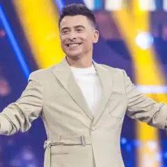 Uśmiechnięty mężczyzna w beżowej marynarce trzyma mikrofon, stoi na tle kolorowej sceny.