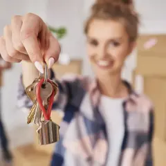 Kobieta trzyma klucze z breloczkiem w kształcie domu, na tle kartonów i mężczyzny.