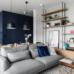 Szara sofa w małym salonie z półkami, stołem i aneksem kuchennym w tle.