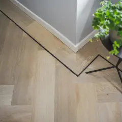 Drewniana podłoga w jodełkę z czarną, geometryczną wstawką. W rogu stoi doniczka z zieloną rośliną, a obok czarny stolik.