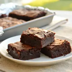 Deliciosos trozos de brownie sin gluten apilados en un plato blanco, con más en un molde al fondo.