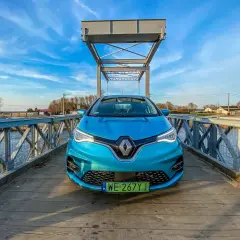 Niebieski Renault Zoe stoi na drewnianym moście nad wodą, z żaglówkami w tle.