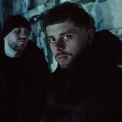 Dwóch mężczyzn w ciemnych kurtkach i czapce na tle zniszczonej ściany.
