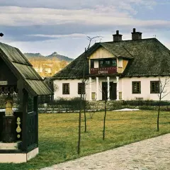 Drewniana kapliczka z krzyżem i figurą, obok restauracja "Dwór". W tle sosny i zabudowania. Idealne miejsce na camping.