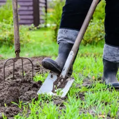 Osoba w gumowcach i czarnych spodniach zaczyna przekopać ogródek, używając łopaty i widł.