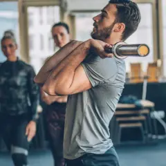 Mężczyzna ćwiczy z sztangą, przygotowując się do FBW Sakera. W tle dwie kobiety obserwują trening.
