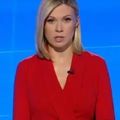 Monika Sawka w czerwonej sukience, z mikrofonem przy dekolcie, na tle niebieskiego ekranu.