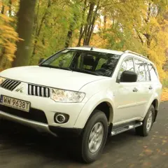 Biały model terenówki Mitsubishi Pajero Sport jedzie po drodze wśród jesiennych drzew.