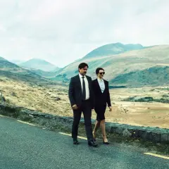 Coppia in abiti eleganti cammina su strada panoramica, evocando il significato di "The Lobster" e la ricerca di un legame in un paesaggio desolato.