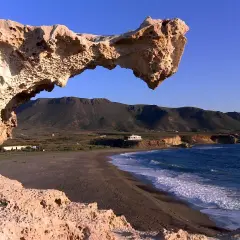 Un arco natural de roca erosionada, como un gigante dormido, vigila una playa solitaria y el mar azul. Los escullos forman un portal a un paisaje sereno.