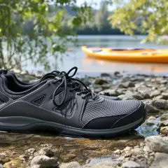 Szare buty sportowe na kamienistej plaży, idealne jako buty na kajaki. W tle żółty kajak na wodzie.