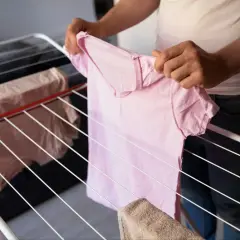 Suszenie prania w domu może zwiększać wilgoć. Różowa koszulka wisi na suszarce obok innych ubrań.