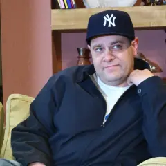 Mężczyzna w czapce z daszkiem Yankees, w wieku, który można by określić jako dojrzały, siedzi na kanapie.
