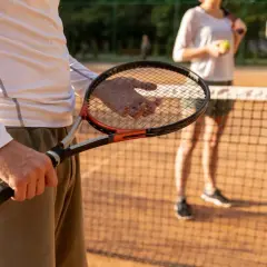Mężczyzna trzyma rakietę tenisową z naciągiem, gotowy do gry na korcie. W tle kobieta z piłką.