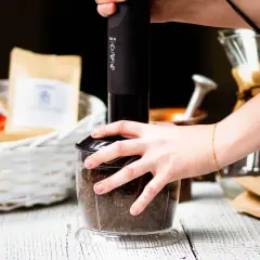 Ręce mielą kawę w blenderze. W tle koszyk z paczkami kawy i dripper.