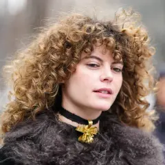 Eine Frau mit voluminösen, krausen Haaren trägt einen Pelzmantel und eine Halskette mit einer goldenen Schleife.