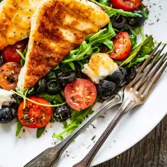 Pyszna sałatka z grillowanym halloumi, pomidorkami koktajlowymi i czarnymi oliwkami na rukoli. Idealna na lekki posiłek.