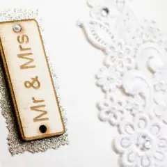 Dettagli di un invito di nozze: targa in legno "Mr & Mrs", pizzo bianco e logo "Party Partenze". Un esempio di diversi tipi di nozze.