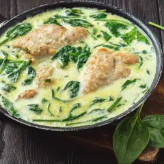 Pyszny kurczak ze szpinakiem w kremowym sosie, podany na patelni. Obok świeże liście szpinaku i czosnek.