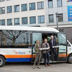 Drei Personen stehen vor einem weißen Kleinbus mit der Aufschrift "WI Mobil". Sie halten Schilder mit der Aufschrift "On-Demand-Shuttle".