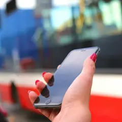 Dłoń z czerwonymi paznokciami trzyma smartfon, na którym można kupić bilety ZTM Warszawa. W tle widać autobus.