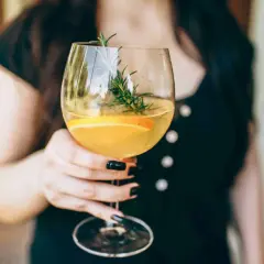 En person håller ett glas med en uppfriskande drink med vitt vin, garnerad med en apelsinskiva och en rosmarinkvist.
