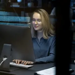 Młoda kobieta w okularach pracuje przy komputerze, na ekranie widać kod. To przyszłość cyberbezpieczeństwa, studia w tej dziedzinie otwierają wiele możliwości.