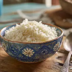 En skål med fluffigt, nykokt ris står på ett rustikt träbord. Bredvid skålen ligger en sked. Perfekt för att koka ris till middagen.