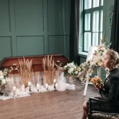 Smutna kobieta z kwiatami siedzi obok trumny. W kulturze nie wkłada się zmarłemu pieniędzy.