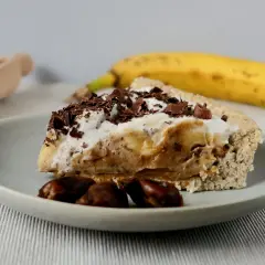 Kawałek pysznego banoffee z bitą śmietaną i czekoladowymi wiórkami, obok dojrzały banan i kilka daktyli.