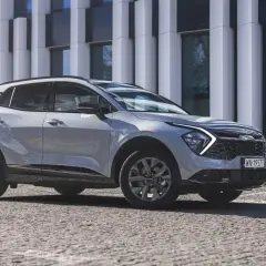 Nowy Kia Sportage Anniversary w jasnym kolorze, zaparkowany przed nowoczesnym budynkiem. Stylowy SUV na kamienistej nawierzchni.