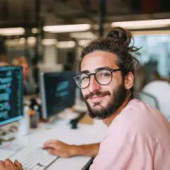 Ein junger Mann mit Brille und Dutt sitzt vor einem Computer, auf dem Code zu sehen ist. Er studiert Informatik an der Fernuni und lächelt in die Kamera.