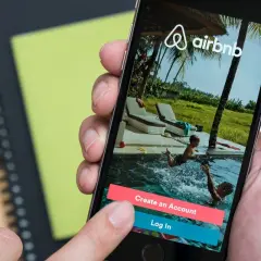 Dłoń trzyma smartfon z aplikacją Airbnb. Widok na basen z dziećmi. Zastanawiasz się airbnb co to? To platforma do rezerwacji noclegów.