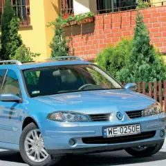 Błękitne kombi Renault Laguna 2 z relingami dachowymi stoi przed domem z żółtą elewacją i drewnianym płotem.